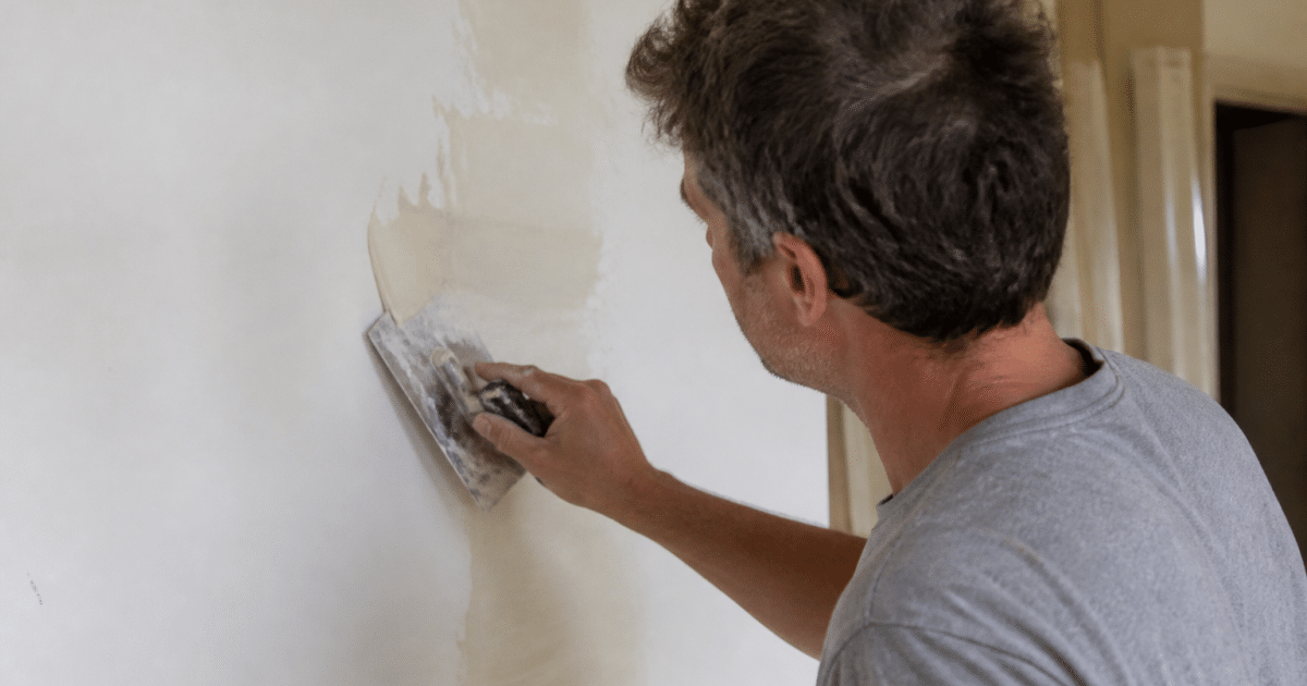main appliquant enduit beige clair sur mur blanc avec spatule